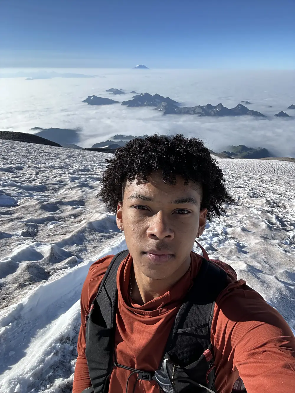 Evan in an orange shirt and pack on a high snowfield, above a sea of clouds with peaks breaking through and a distant volcano on the horizon.